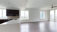 This single-floor space showcases a thoughtfully designed open-concept layout that seamlessly integrates the kitchen and living areas into one cohesive environment. Rich, dark hardwood flooring extends throughout, adding warmth and contrast against the crisp white walls and ceilings. Recessed lighting is installed overhead, providing even, unobtrusive illumination across the space.

The kitchen is outfitted with dark espresso cabinetry, a neutral tile backsplash, and a clean white countertop that extends into a peninsula with an integrated sink — offering both functional workspace and a subtle visual boundary between the kitchen and living area. The modern faucet adds a refined finishing touch.

Large sliding windows dominate the exterior walls, flooding the interior with natural light and framing impressive views of a city skyline and surrounding greenery. The living area benefits from this abundant natural light, creating an airy, open ambiance. A second window on the adjacent wall further enhances ventilation and views of neighboring modern architecture. The overall aesthetic is clean, minimalist, and well-suited to contemporary urban living.