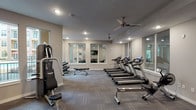 This single-floor fitness center is thoughtfully designed to support a wide range of workout routines. The space is divided into two primary areas: a large main gym floor and a separate studio room, connected by glass-paneled doors that maintain an open, cohesive feel throughout.

The main workout area is generously sized and features patterned carpet tile flooring in neutral gray tones. Multiple ceiling fans and recessed lighting ensure consistent airflow and visibility throughout. The cardio zone includes a row of treadmills and elliptical machines, each equipped with individual display screens. Strength training is well-represented with cable machine systems, a power tower, a leg press, weight benches, and a dumbbell rack. Large mirrors line portions of the walls, enhancing the sense of space. Windows along multiple walls invite natural light and offer views of the surrounding property.

The dedicated studio room is a standout feature, offering a clean, open layout ideal for stretching, yoga, or group fitness. It is lined with floor-to-ceiling mirrors on one wall, complemented by a ceiling fan, recessed lighting, and a window that brings in natural light and courtyard views.

Overall, the fitness center presents a modern, clean aesthetic with neutral walls, consistent flooring, and a practical layout that efficiently accommodates both cardio and strength training needs.