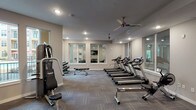 This single-floor fitness center is thoughtfully designed to support a wide range of workout routines. The space is divided into two primary areas: a large main gym floor and a separate studio room, connected by glass-paneled doors that maintain an open, cohesive feel throughout.

The main workout area is generously sized and features patterned carpet tile flooring in neutral gray tones. Multiple ceiling fans and recessed lighting ensure consistent airflow and visibility throughout. The cardio zone includes a row of treadmills and elliptical machines, each equipped with individual display screens. Strength training is well-represented with cable machine systems, a power tower, a leg press, weight benches, and a dumbbell rack. Large mirrors line portions of the walls, enhancing the sense of space. Windows along multiple walls invite natural light and offer views of the surrounding property.

The dedicated studio room is a standout feature, offering a clean, open layout ideal for stretching, yoga, or group fitness. It is lined with floor-to-ceiling mirrors on one wall, complemented by a ceiling fan, recessed lighting, and a window that brings in natural light and courtyard views.

Overall, the fitness center presents a modern, clean aesthetic with neutral walls, consistent flooring, and a practical layout that efficiently accommodates both cardio and strength training needs.