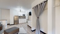 Located at 1255 S SH 6 Access Rd in Houston, TX, this single-floor studio unit offers a thoughtfully designed open-concept layout that maximizes functionality within a compact footprint. Light-toned luxury vinyl plank flooring runs throughout, lending a cohesive and contemporary feel to the entire space.

The living area is bright and welcoming, featuring crisp white walls, a textured ceiling, and natural light provided by a window fitted with a roller shade. A plush gray sofa with a matching ottoman anchors the space, complemented by a white nightstand and a wall-mounted flat-screen TV.

Transitioning seamlessly from the living area, the kitchenette is well-appointed with white shaker-style cabinetry, a mosaic tile backsplash in neutral tones, light countertops, and stainless steel appliances including a full-size refrigerator and a built-in microwave. A single-basin sink and compact cooktop complete the functional cooking space. A sputnik-style chandelier adds a modern design accent overhead.

A curtained closet area with a built-in shelf provides convenient storage, while a white dresser offers additional organization. The bathroom is clean and efficiently laid out, featuring a marble-look countertop with an integrated sink, a large mirror with cabinet, and wood-look flooring that ties back to the rest of the unit.