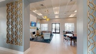 This thoughtfully designed single-floor clubhouse offers a full suite of amenities tailored for community gathering, fitness, and leisure. The entrance sets a refined tone with soaring ceilings, herringbone-patterned hardwood flooring, and a striking sculptural gold chandelier framing the glass entry doors. The main lounge area is anchored by a floor-to-ceiling white brick fireplace with a wood mantel, a wall-mounted TV, and comfortable seating arranged on a patterned area rug — all beneath a coffered ceiling with warm pendant lighting and large windows that invite abundant natural light. Adjacent to the lounge, a well-appointed kitchen features light wood cabinetry, a white quartz island, pendant lighting, a stylized open shelving display, and modern appliances, flowing seamlessly into a dining area with round tables and teal accent chairs. A dedicated game room offers shuffleboard, foosball, plush seating, and natural window views — ideal for social interaction. The fitness facilities include a fully equipped strength and cardio gym with multiple machines and wall-mounted TVs, as well as a separate cycling studio with spin bikes, herringbone hardwood floors, and curated wall art. A water fountain station and accessible restrooms provide convenience throughout. The overall aesthetic is contemporary and cohesive, combining neutral tones with teal accents and warm wood elements.