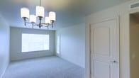 This single-floor space presents a clean, well-lit interior defined by soft, neutral tones throughout. The main room is finished with plush wall-to-wall carpeting in a light gray hue, contributing to a calm and comfortable atmosphere. Walls are painted in a muted blue-gray shade, lending the space a serene, contemporary feel.

A standout feature of the room is the chandelier, which showcases a brushed nickel finish with cylindrical white shades arranged in a classic multi-arm configuration, adding a refined focal point to the ceiling. Natural light enters generously through a large window, brightening the interior and creating an airy, open ambiance.

The room includes a paneled interior door with an arched top detail, finished in crisp white, which adds a traditional architectural element to the otherwise modern-neutral design. A secondary glass-paneled door is also visible, likely providing access to an exterior area and further enhancing the flow of natural light.

Overall, the space offers a bright, move-in-ready interior with thoughtful finishes that balance classic and contemporary design elements.