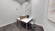 Located at 285 Park Ave in Savannah, Georgia, this single-floor workspace offers a thoughtfully designed and functional environment suited for focused, professional work. The space features a shared workstation setup with a sleek white dual-sided desk equipped with desktop computers and a natural wood privacy divider, providing a degree of separation between workstations while maintaining an open, collaborative feel.

The interior is finished in soft sage-green walls that lend a calm, neutral ambiance throughout. Dark hardwood-style flooring contrasts cleanly with the light wall tones, adding warmth and grounding the overall aesthetic. Modern chrome-framed rolling chairs complement the contemporary design language of the furnishings.

A decorative wall panel treatment serves as a focal backdrop, displaying two gold-framed landscape artworks stacked vertically, adding a refined artistic element to the space. The clean white baseboards and recessed panel detailing on the accent wall contribute to the polished, professional finish. The layout is efficiently organized, making practical use of the available footprint while maintaining an uncluttered, orderly atmosphere conducive to productivity.
