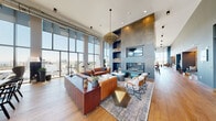The Club Room at The Smyth is an expansive, single-floor amenity space that combines upscale design with versatile social functionality. Dramatic floor-to-ceiling windows wrap the perimeter, flooding the interior with natural light and offering sweeping panoramic views of the surrounding cityscape. Wide-plank hardwood flooring runs throughout, grounding the space with warmth and continuity.

The lounge area anchors one end of the room, featuring a striking floor-to-ceiling slate-finished fireplace surround with built-in shelving, a wall-mounted TV, and a grand chandelier overhead. Richly upholstered leather sofas, eclectic accent chairs, and layered area rugs create an inviting, hotel-inspired atmosphere. A large communal dining table positioned near expansive glass walls serves as an ideal gathering spot bathed in natural light.

The entertainment zone includes a full-size billiards table, a shuffleboard table, and a stylish wet bar with a dark navy island, leather-backed barstools, and textured tile backsplash. Overhead industrial-style shelving adorned with greenery adds character. Teal channel-tufted booth seating provides intimate dining or lounge nooks. A dedicated catering kitchen with sleek cabinetry and quartz countertops offers practical support for events. Cozy reading alcoves with velvet armchairs complete this thoughtfully designed communal retreat.