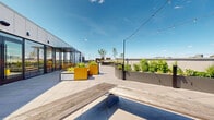 Located at 321 Main St in Hackensack, NJ, this expansive rooftop terrace offers a thoughtfully designed outdoor experience with multiple distinct zones for gathering, dining, and relaxing. The terrace features large-format porcelain tile flooring throughout most of the space, transitioning to warm wood deck panels in select areas, creating visual variety underfoot. String lights are strung across the entire rooftop, adding ambient evening appeal.

A central pergola structure anchors the entertaining zone, housing a built-in outdoor bar with bar-height seating, ideal for social gatherings. Adjacent to this, a substantial communal dining table with bench seating provides a focal point for group events. Multiple white lounge chairs, sofas, and bistro-style tables are distributed across the terrace to accommodate various group sizes.

Raised planters line the perimeter, filled with lush greenery including ornamental grasses, shrubs, and young trees, complemented by horizontal wood-slat privacy screens. Large floor-to-ceiling glass doors and windows connect the terrace to interior spaces, allowing seamless indoor-outdoor flow and abundant natural light. The rooftop commands open views of the Hackensack skyline and surrounding urban landscape, with the New York City skyline visible on the horizon, making it a standout feature of this property.