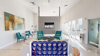 Located in Scottsdale, Arizona, this dynamic community amenity space is designed for socializing and recreation. The open-plan layout is anchored by soaring ceilings that amplify the sense of space, while expansive floor-to-ceiling glass doors and windows flood the interior with abundant natural light and provide views of the surrounding Scottsdale landscape.

The lounge area serves as the social heart of the room, featuring a neutral-toned sofa and teal accent chairs arranged around a sleek round coffee table atop a chartreuse area rug. Vibrant abstract artwork adorns the crisp white walls, adding pops of color throughout. A bold horizontal-slat accent wall houses a wall-mounted flat-screen TV, creating a striking focal point that divides the lounge from the billiards area.

The billiards zone is centered around a wood-framed pool table with teal felt, positioned to take full advantage of the natural light pouring in from multiple glass doors that open directly to the outdoors. High-top tables with bar stools offer additional casual seating nearby. A giant Connect Four game adds a playful, interactive element to the space.

The polished concrete flooring ties the entire area together, contributing to the modern, clean aesthetic that defines this well-appointed Scottsdale amenity space.