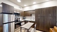 This thoughtfully designed single-floor studio offers a seamless open layout that integrates the kitchen, living, and sleeping areas into one cohesive space. The kitchen is a standout feature, showcasing rich dark walnut-toned cabinetry that extends floor to ceiling for ample storage, complemented by crisp white countertops and a full suite of stainless steel appliances, including a refrigerator, electric range, over-the-range microwave, and dishwasher. Recessed ceiling lights illuminate the kitchen evenly, while a compact dining table with chairs provides a practical eating area. Light hardwood flooring flows throughout the entire unit, lending warmth and continuity. The living and sleeping area is bright and open, anchored by a large window that welcomes natural light and leafy outdoor views. A wall-mounted flat-screen television, a leather accent chair, and a full-size bed make efficient use of the space. The bathroom features clean, minimalist finishes, with a white quartz vanity countertop, an undermount sink, brushed chrome faucet, and a large mirror. The overall aesthetic is modern and functional, with a neutral color palette and contemporary design elements throughout.