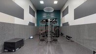 This single-floor space is thoughtfully designed to serve as a multifunctional amenity area, combining a professional-grade fitness center with a well-appointed kitchen and lounge area. The fitness center features a bold, energizing aesthetic with teal accent walls, motivational signage, and graphic gray-and-white striped upper walls. Rubber flooring provides a durable workout surface, and the space is outfitted with a functional trainer cable machine, a heavy punching bag, kettlebells, a BOSU ball, a flat bench, and battle ropes, along with a sculptural pendant light overhead. Recessed panel lighting illuminates the room efficiently. The kitchen area is finished with rich, dark wood cabinetry extending to the ceiling, light granite countertops, and neutral tile flooring. Stainless steel appliances include a full-size refrigerator, a dishwasher, an electric range with a stainless steel backsplash, and a microwave. A deep sink and ample counter space add practicality. An adjacent open area features a large window overlooking the surrounding neighborhood, allowing natural light to fill the space. A suspended drop-tile ceiling and clean neutral walls complete the polished, functional interior throughout.