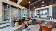 This contemporary single-floor commercial space is thoughtfully designed to balance functionality with a welcoming, upscale aesthetic. Upon entry, polished tile flooring transitions into carpeted lounge areas, creating defined zones throughout the open layout. Large floor-to-ceiling windows along the street-facing wall invite abundant natural light and offer views of the surrounding streetscape, enhancing the airy atmosphere of the space.

The main lounge area is furnished with a mix of round stainless steel bistro tables paired with bentwood chairs, as well as cushioned lounge seating and decorative accent stools. A prominent dark wood-paneled accent wall anchors a built-in service counter with cabinetry below and a large flat-screen television mounted above, serving as a central focal point. Pendant lighting and an ornate chandelier add layers of illumination against the dramatic exposed-duct black ceiling.

A glass-walled conference or private meeting room is visible from the main area, offering transparency while maintaining a sense of separation. It features a long dining-style table with upholstered chairs and framed artwork. Additional seating nooks with armchairs and side tables provide quieter areas for focused use. Vibrant abstract artwork punctuates the walls throughout, contributing energy and color to the overall sophisticated, modern ambiance.