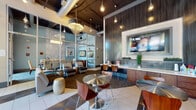 This contemporary single-floor commercial space is thoughtfully designed to balance functionality with a welcoming, upscale aesthetic. Upon entry, polished tile flooring transitions into carpeted lounge areas, creating defined zones throughout the open layout. Large floor-to-ceiling windows along the street-facing wall invite abundant natural light and offer views of the surrounding streetscape, enhancing the airy atmosphere of the space.

The main lounge area is furnished with a mix of round stainless steel bistro tables paired with bentwood chairs, as well as cushioned lounge seating and decorative accent stools. A prominent dark wood-paneled accent wall anchors a built-in service counter with cabinetry below and a large flat-screen television mounted above, serving as a central focal point. Pendant lighting and an ornate chandelier add layers of illumination against the dramatic exposed-duct black ceiling.

A glass-walled conference or private meeting room is visible from the main area, offering transparency while maintaining a sense of separation. It features a long dining-style table with upholstered chairs and framed artwork. Additional seating nooks with armchairs and side tables provide quieter areas for focused use. Vibrant abstract artwork punctuates the walls throughout, contributing energy and color to the overall sophisticated, modern ambiance.