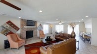 This single-floor residence offers a well-designed open-concept layout that seamlessly connects the living and dining areas, creating a sense of generous, flowing space. The living room serves as the heart of the home, anchored by a full-height brick fireplace with a classic white mantel and a wall-mounted television above, providing both a functional and aesthetic focal point. Rich brown leather sofas are arranged around a wooden coffee table atop a bold, patterned area rug, lending the space a warm and welcoming atmosphere. Recessed lighting and a ceiling fan keep the room bright and comfortable throughout the day. Large windows along the perimeter allow abundant natural light to fill the interior, enhancing the airy feel of the space. The adjacent dining area is visible from the living room, featuring a dining table set beneath additional natural light sources, maintaining the open and connected character of the floor plan. The flooring throughout appears to be light-toned tile, contributing to a clean and cohesive look. A textured ceiling adds subtle architectural detail, completing the overall inviting and practical character of this single-level home.