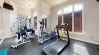 Located at 3131 Simpson Stuart Rd in Dallas, TX, this dedicated fitness center offers a well-appointed exercise space designed to support a full range of workout routines. The room features elegant architectural details, including crown molding, elevated ceilings, and large arched windows dressed with wood-toned blinds that allow generous natural light to fill the space. The walls are finished in a warm neutral tone, complemented by white baseboards and trim that lend the room a clean, polished appearance. Dark rubber flooring provides a durable, slip-resistant surface appropriate for both cardio and strength training activities. A ceiling fan overhead contributes to comfortable ventilation during workouts. The room is equipped with a treadmill, an elliptical cross-trainer, and a multi-station cable weight machine, providing users with a comprehensive set of cardio and resistance training options. Full-length wall mirrors amplify the sense of space and allow users to monitor their form. A wall-mounted flat-screen television adds an entertainment option during exercise sessions. The fitness center is accessed through white-framed French doors that connect to an adjoining corridor, and posted fitness rules near the entrance help ensure shared use of the space remains orderly and enjoyable for all users.