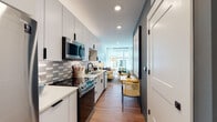 This single-floor residence is thoughtfully designed to maximize functionality while maintaining a clean, modern aesthetic throughout. The galley-style kitchen runs along one side of the open layout and is outfitted with stainless steel appliances — including an electric range, over-the-range microwave, and dishwasher — complemented by crisp white cabinetry with matte black hardware, light quartz countertops, and a stylish geometric tile backsplash in muted silver and gray tones. Warm hardwood-style flooring flows continuously through the space, creating cohesion from the kitchen through the dining and living areas.

A compact dining nook sits adjacent to the kitchen, followed by an open living area that benefits from a large window allowing natural light to fill the space. A partial wall divides the living area from the sleeping zone, which features its own window and ample room for a full bedroom setup.

The bathroom is finished with large-format white wall tiles, wood-toned flooring, a white vanity, and matte black fixtures. Direct access to a walk-in closet with built-in shelving and a stacked washer and dryer adds significant convenience. Recessed lighting and soft blue-gray accent walls contribute to the space's calm, contemporary ambiance.