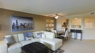 This single-floor space showcases an open-concept layout that seamlessly connects the living, dining, and kitchen areas into one cohesive and functional environment. The living area is anchored by a generous cream-colored sectional sofa paired with decorative throw pillows, complemented by a tufted dark leather ottoman that serves as a striking focal point. A ceiling fan with integrated lighting is centrally positioned overhead, ensuring both comfort and illumination throughout the space. Warm taupe-toned walls create an inviting atmosphere, accented by a large cityscape artwork that adds a contemporary urban character to the room. A trio of mirrored wall panels enhances the sense of depth and reflects natural light effectively. The dining area accommodates a round dark wood table with white upholstered chairs, ideal for intimate gatherings. Beyond the dining space, the kitchen features white cabinetry, open shelving, and essential appliances, maintaining the clean and modern aesthetic carried throughout. Neutral-toned carpeting grounds the space, adding warmth underfoot, while a geometric patterned area rug defines the seating zone. Overall, the space presents a polished, contemporary ambiance suitable for comfortable everyday living.