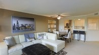 This single-floor space showcases an open-concept layout that seamlessly connects the living, dining, and kitchen areas into one cohesive and functional environment. The living area is anchored by a generous cream-colored sectional sofa paired with decorative throw pillows, complemented by a tufted dark leather ottoman that serves as a striking focal point. A ceiling fan with integrated lighting is centrally positioned overhead, ensuring both comfort and illumination throughout the space. Warm taupe-toned walls create an inviting atmosphere, accented by a large cityscape artwork that adds a contemporary urban character to the room. A trio of mirrored wall panels enhances the sense of depth and reflects natural light effectively. The dining area accommodates a round dark wood table with white upholstered chairs, ideal for intimate gatherings. Beyond the dining space, the kitchen features white cabinetry, open shelving, and essential appliances, maintaining the clean and modern aesthetic carried throughout. Neutral-toned carpeting grounds the space, adding warmth underfoot, while a geometric patterned area rug defines the seating zone. Overall, the space presents a polished, contemporary ambiance suitable for comfortable everyday living.