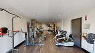 Located in Peoria, Arizona, this fitness facility offers a single, elongated exercise room designed to accommodate a comprehensive range of workout activities. The space features wood-look plank flooring that runs the full length of the room, complemented by neutral-toned walls and recessed ceiling lighting that creates a bright, focused atmosphere throughout.

The room is well-appointed with a diverse selection of fitness equipment. Cardio options include treadmills, elliptical trainers, a rowing machine, and a recumbent cycling unit, providing users with ample variety for aerobic training. Strength training is equally supported, with a multi-station cable machine, a weight bench, a dumbbell rack with free weights, and additional resistance equipment. A wall-mounted flat-screen television is positioned at one end of the room, enhancing the workout experience.

Natural light enters through glass-panel doors at one end, offering a view to the exterior and contributing to the room's open, airy feel. The layout is spacious enough to allow comfortable movement between equipment stations. Additional functional elements include visible HVAC vents, an emergency exit sign, and a fire extinguisher, reflecting attention to safety and comfort. The overall design is clean, practical, and well-suited for a broad range of fitness routines.