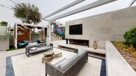 Located at 1961 E Dyer Rd in Santa Ana, California, this thoughtfully designed single-level outdoor space delivers a resort-inspired experience with a variety of distinct amenity zones. At its heart is a sparkling pool surrounded by a clean concrete deck, accented by patches of artificial turf and unique woven hanging lounge chairs that invite relaxation. String lights overhead lend a warm, inviting ambiance during evening hours, while tropical plantings — including banana palms and lush greenery — create a verdant backdrop.

A covered outdoor lounge area anchors one end of the space, featuring a sleek linear fireplace set into a floor-to-ceiling stone tile wall, a wall-mounted television, and ample seating with wicker and upholstered furnishings arranged over a geometric-patterned area rug. The pergola-style overhead structure frames open sky views while providing comfortable shade.

A striking artistic centerpiece — a large-scale tropical mural by artist Carly Ealey — wraps an architectural structure near the stairway, adding bold color and creative energy to the environment. Wide-step staircases with chevron-patterned tile connect the different levels of the space elegantly. Thoughtfully curated landscaping throughout features agaves, succulents, sculpted shrubs, and mature trees, completing this well-appointed outdoor destination.