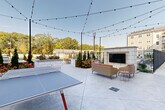 This thoughtfully designed outdoor courtyard in Peabody, Massachusetts offers a comprehensive array of amenities within a beautifully landscaped setting. At the heart of the space is a sparkling resort-style swimming pool, surrounded by a spacious sundeck lined with lounge chairs, ideal for relaxing in the open air. Adjacent to the pool area, a fully equipped outdoor kitchen features a stainless steel gas grill and tiled countertops, perfect for al fresco cooking and entertaining. A dedicated lounge area centers around a stone fireplace fitted with a large outdoor television, furnished with comfortable wicker seating and accented by string lights that create an inviting evening atmosphere. A ping-pong table adds a recreational element to the patio space. Freestanding hammocks are nestled beside curated garden beds, providing a serene retreat surrounded by evergreen plantings and seasonal flowers. Manicured lawns extend throughout the courtyard, complemented by winding concrete pathways and thoughtful landscaping. Rocking chairs and accent seating are positioned along the building's exterior, offering quiet nooks to unwind. The multi-story building surrounding the courtyard features private balconies, large windows, and classic lap siding, framing the outdoor space with a polished residential character.