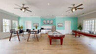 Located at 2 Abbott Drive in Hampton, Virginia, this single-floor community clubhouse offers a well-appointed and versatile gathering space designed for both recreation and functionality. The interior is characterized by light hardwood flooring, crisp crown molding, and recessed lighting throughout, creating a clean and polished aesthetic.

The main living and dining area is a standout feature, adorned with bold aqua-toned walls accented by white wainscoting panels. High-top wooden dining tables with modern mesh-back bar stools provide ample seating, while a kitchenette with granite countertops and stainless steel appliances adds convenience for entertaining.

An adjacent game room opens through large floor-to-ceiling glass doors to an outdoor pool area, flooding the space with natural light and garden views. This room houses a pool table and a shuffleboard table beneath a ceiling fan, making it an ideal space for social activities and leisure.

The reception area features soft gray walls, a built-in granite-topped workstation with white cabinetry, a wall-mounted flat-screen television, and French doors that overlook lush greenery. Framed artwork is displayed throughout the space, adding a refined, curated touch to the overall ambiance. The clubhouse seamlessly blends comfort, style, and community-oriented amenities.