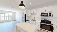 This thoughtfully designed one-bedroom, one-bathroom unit offers a clean, modern aesthetic across a single floor. The open-concept kitchen serves as a standout feature, showcasing crisp white shaker cabinetry with matte black hardware, a full suite of stainless steel appliances including a refrigerator, electric range, built-in microwave, and dishwasher, as well as a single-basin sink with a black faucet and light quartz countertops. A freestanding white island with lower shelving provides additional prep space and storage, while a bold black pendant light anchors the space above. Light wood-style flooring flows throughout the kitchen and living areas, contributing to a bright, airy ambiance. The living room benefits from a large window that invites natural light into the space. The bedroom is carpeted for comfort and features a sizable window with views of the surrounding building exterior, allowing ample daylight. A dedicated walk-in closet with wire shelving and hanging rods provides generous storage. The bathroom is finished with a full tub-shower combination, white surround, and matte black fixtures, maintaining the consistent modern design language found throughout the unit. Overall, the space presents a cohesive, contemporary style with a neutral palette.