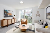 This contemporary single-floor residence offers a well-organized layout that balances comfort and modern design. An inviting entryway corridor, accented by a round gold-framed mirror and recessed lighting, leads into the heart of the unit. The open-concept living area is bathed in natural light from large floor-to-ceiling windows that frame views of lush greenery and the surrounding neighborhood, creating a bright and airy atmosphere. The living space is thoughtfully arranged with room for seating and lounge furniture, complemented by curated artwork on the walls. Adjacent to the living area, the kitchen is a functional and stylish centerpiece, featuring light wood cabinetry with matte black hardware, stone-look countertops, a stainless steel undermount sink with a matte black faucet, and a striking herringbone-patterned tile backsplash. The bedroom offers a serene retreat with wood-look flooring, a ceiling fan with integrated lighting, a wall-mounted sconce, and an open wire-shelving closet for organized storage. The bathroom is clean and practical, finished with warm-toned flooring, a wood vanity with stone countertop, and ample space. Throughout the unit, light-toned walls, recessed lighting, and cohesive modern finishes create a harmonious and welcoming environment.