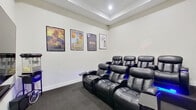 This dedicated home theater room is designed to deliver an immersive cinematic experience. The space is arranged with two tiers of plush black leather power recliner seats, each equipped with built-in cup holders, swivel tray tables, and glowing blue LED accent lighting along the bases—adding a dramatic, theater-like ambiance. The seating is positioned to face a large flat-screen television mounted on the front wall, complemented by a subwoofer for enhanced audio performance.

The room features neutral white walls adorned with an extensive collection of framed classic movie posters, including titles such as Spider-Man, The Karate Kid, Dirty Dancing, Bohemian Rhapsody, A Star Is Born, and Back to the Future, lending the space a curated, cinematic personality. A vintage-style popcorn machine on a wheeled cart adds a fun, authentic movie-house touch.

The ceiling incorporates a recessed tray detail with centered lighting, adding architectural depth to the room. Dark carpet throughout absorbs sound and enhances the overall theater aesthetic. A paneled white entry door with a brass kick plate provides access to the space, completing this well-appointed, single-floor entertainment room.