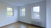 This single-floor space presents a clean, minimalist aesthetic defined by crisp white walls and rich hardwood flooring that runs throughout. The room is notably well-lit, benefiting from multiple double-hung windows that frame views of surrounding trees and the outdoors, filling the interior with generous amounts of natural light. The windows, trimmed in white, complement the neutral palette of the space and contribute to an airy, open atmosphere. A classic cast-iron radiator positioned beneath one of the windows serves as both a functional heating element and a charming architectural detail reminiscent of traditional urban residential design. The white baseboards provide clean definition along the floor line, adding to the overall sense of order and simplicity. The ceiling height appears comfortable and proportional, reinforcing the room's open feel. The hardwood floors display warm, natural tones with character that adds depth and texture to the otherwise understated interior. Overall, the space offers a bright, well-maintained environment with timeless details, making it a versatile and inviting setting suited to a variety of uses.