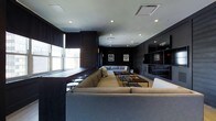This single-floor space exudes a sleek, contemporary ambiance anchored by a bold, dark color palette of deep charcoal and rich wood tones. The main lounge area is generously proportioned, designed for comfort and social gathering, and is furnished with large, plush sectional sofas in neutral gray tones arranged around low-profile wooden coffee tables — ideal for relaxed conversation or entertainment. Recessed ceiling lighting provides a clean, even illumination throughout, complementing the modern aesthetic. A row of windows along one wall invites natural light and a connection to the outside, while roller shades offer privacy and light control. The dark wall paneling lends a refined, club-like atmosphere, further enhanced by framed sports jerseys displayed as wall art, adding a distinctive personality to the space. A built-in shelving and media unit along the far wall provides both functional storage and a striking architectural focal point. Bar-height seating along the window ledge offers an additional casual seating option. Overall, the space strikes a balance between a high-end lounge and a sports entertainment venue, making it well-suited for communal gatherings and social events.