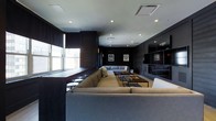 This single-floor space exudes a sleek, contemporary ambiance anchored by a bold, dark color palette of deep charcoal and rich wood tones. The main lounge area is generously proportioned, designed for comfort and social gathering, and is furnished with large, plush sectional sofas in neutral gray tones arranged around low-profile wooden coffee tables — ideal for relaxed conversation or entertainment. Recessed ceiling lighting provides a clean, even illumination throughout, complementing the modern aesthetic. A row of windows along one wall invites natural light and a connection to the outside, while roller shades offer privacy and light control. The dark wall paneling lends a refined, club-like atmosphere, further enhanced by framed sports jerseys displayed as wall art, adding a distinctive personality to the space. A built-in shelving and media unit along the far wall provides both functional storage and a striking architectural focal point. Bar-height seating along the window ledge offers an additional casual seating option. Overall, the space strikes a balance between a high-end lounge and a sports entertainment venue, making it well-suited for communal gatherings and social events.
