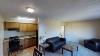 This single-floor unit offers a practical and functional layout that combines the kitchen and living area in one open space. The kitchen is situated along one wall, featuring warm wood-toned cabinetry, a double-basin sink, and a black range, all set over light hardwood-style flooring. Upper and lower cabinets provide ample storage, and a fluorescent light fixture ensures the workspace is well illuminated.

The living area flows naturally from the kitchen, transitioning to carpeted flooring. A dark leather sofa serves as the focal point of the sitting space, complemented by two matching end tables and a larger coffee table, all in a dark wood finish. A pair of table lamps adds warm ambient lighting to the room. The textured popcorn ceiling runs throughout the unit, and flush-mounted ceiling lights provide general illumination.

A doorway visible from the living area leads to a separate room, suggesting a private bedroom or additional space. The neutral, light-colored walls and straightforward layout give the unit a clean, no-frills character suited for practical, everyday living.