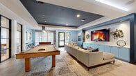 Located at 881 W Warren Ave in Longwood, Florida, this contemporary community lounge and game room offers a well-appointed single-floor space designed for socializing and recreation. The room is defined by its striking tray ceiling with a bold dark finish, which contrasts elegantly against the soft sky-blue walls and crisp white trim throughout.

The entertainment amenities are a clear highlight, featuring a full-size pool table as a central focal point, a shuffleboard table, and a foosball table, providing a diverse range of recreational options. A large flat-screen TV is mounted within a built-in display wall flanked by custom light-blue cabinetry and decorative metallic wall art, creating a polished media and lounge area. A generously sized sectional sofa anchors this viewing zone atop a layered area rug.

Large floor-to-ceiling windows and glass-panel doors invite ample natural light and offer views of the surrounding outdoor greenery. Large-format tile flooring adds a clean, modern foundation throughout the space. Curated abstract artwork adorns the walls, enhancing the upscale aesthetic. A glazed partition connects this lounge to an adjacent room, further expanding the sense of openness. The overall ambiance is modern, vibrant, and thoughtfully designed for community engagement.