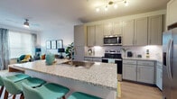 This thoughtfully designed single-floor residence offers a cohesive open-concept layout that seamlessly connects the kitchen, dining, and living areas. The kitchen is a standout feature, boasting shaker-style grey cabinetry that extends to the ceiling for ample storage, a full suite of stainless steel appliances including a French door refrigerator, electric range, and built-in microwave, all complemented by a white subway tile backsplash. A generous island with granite countertops and an undermount sink serves as both a functional prep area and a casual dining spot with seating for several. Light wood-style flooring flows throughout the main living areas, enhancing the bright and airy atmosphere. The living room receives natural light through a large window dressed with patterned curtains, creating a warm and inviting retreat. The bedroom continues the vibrant, well-lit aesthetic with a sizable window, ample wall space, and a cozy reading nook. The bathroom features granite-topped grey vanity cabinetry, a clean layout with wood-style flooring, and generous hallway storage. Throughout the space, recessed and track lighting ensures every area is well-illuminated, while the consistent use of neutral tones accented with colorful décor creates a modern and welcoming ambiance.