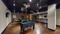 This single-floor entertainment space blends industrial-modern design with a vibrant recreational atmosphere. The open layout is anchored by rich hardwood flooring and a dramatic black exposed ceiling with visible ductwork, track lighting, and ceiling fans that contribute to the room's loft-like character.

The game area serves as the centerpiece of the space, housing a full-size pool table with green felt and a foosball table, both positioned to allow ample room for play. A wall-mounted flat-screen television provides additional entertainment options. Along one wall, a stylish bar counter features a stainless steel front panel and a dark wood top, complemented by sleek wire-frame bar stools — ideal for socializing.

A lounge area is defined by tufted yellow leather benches and decorative throw pillows, set against a textured exposed brick wall that adds warmth and character. Tall decorative plants further enhance the inviting atmosphere.

Large floor-to-ceiling glass partition windows along one side of the room provide a visual connection to an adjacent corridor, adding transparency and a sense of depth. Wall art pieces, including a bold red-toned painting, add curated visual interest throughout the space. A large mirror on one wall amplifies the sense of openness. Overall, this space is thoughtfully designed for both leisure and social interaction.