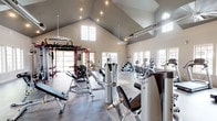 This fitness center is housed within a single-level space defined by dramatic vaulted ceilings that create an open, airy atmosphere throughout. Large gridded windows and floor-to-ceiling glass panels line the walls, flooding the interior with natural light and offering views of the surrounding outdoor areas. The flooring transitions between durable wood-tone and tile surfaces, delineating distinct workout zones across the expansive floor plan.

The strength training area is anchored by a bold red multi-functional cable and functional training rig, complemented by a range of selectorized machines, free weights, kettlebells, and a heavy punching bag. Full-length mirrors along one wall enhance the sense of space and allow users to monitor their form. The cardio section is well-appointed with stair climbers, ellipticals, recumbent bikes, and treadmills positioned to take advantage of natural light from nearby windows.

Additional amenities include a water fountain, built-in storage shelving, and a ceiling fan for air circulation. Recessed and track lighting supplements daylight throughout. A separate entry corridor with glass partitions provides a clean transition into the facility, which maintains a modern, neutral aesthetic with gray and white tones accented by the vivid red equipment framework.