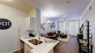 This single-floor residence offers a thoughtfully designed open-concept layout that seamlessly connects the kitchen, dining, and living areas. The kitchen is outfitted with stainless steel appliances, a subway tile backsplash, white cabinetry, and a quartz countertop island with a double-basin sink — providing ample prep and storage space. Adjacent to the kitchen, the dining area features built-in shelving and flows naturally into the living room, which is brightened by large windows with blinds and a ceiling fan. Rich hardwood-style flooring runs throughout the main living areas, adding warmth and continuity. A dedicated laundry area with a full-size washer and dryer is conveniently located near the entryway. The two bedrooms are carpeted for comfort, each equipped with ceiling fans and large windows that allow for generous natural light. The bathroom includes a wide vanity with white cabinetry, quartz countertops, an expansive mirror, and modern pendant lighting, connecting directly to the living spaces via hardwood-style flooring. The overall design maintains a clean, neutral palette with contemporary finishes throughout, creating a cohesive and functional living environment.
