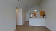 This single-story space showcases an open-concept layout that connects the living area to the kitchen, creating a sense of spaciousness enhanced by vaulted ceilings that draw the eye upward. The living area features neutral beige carpeting, which adds warmth to the light-toned walls and contributes to the overall understated aesthetic of the space.

The kitchen is positioned at the far end of the open area and is defined by a white breakfast bar that serves as both a functional workspace and a natural divider between the kitchen and living spaces. A pendant light hangs above the bar, providing focused illumination. The kitchen is equipped with oak-toned cabinetry offering ample storage, along with standard appliances including a microwave. The white surfaces and light wood tones maintain a clean, cohesive look.

A hallway is visible off the main living area, leading to additional rooms through white paneled doors. Ceiling vents are integrated throughout, ensuring consistent climate control. The overall ambiance of the space is calm and functional, with a neutral color palette and simple finishes that provide a versatile foundation for a variety of interior styles.