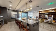 Located at 2000 N Broadway in Denver, Colorado, this sophisticated amenity lounge is a single-floor space offering a generously sized, open-concept layout designed for both socializing and relaxation. The space is divided into several distinct yet interconnected zones, each thoughtfully appointed with upscale finishes and modern furnishings.

The kitchen area is a standout feature, equipped with stainless steel appliances including a full-size refrigerator and built-in oven, sleek flat-panel wood cabinetry, and a marble-style backsplash. A large central island with a polished concrete-style countertop provides ample bar seating beneath a striking sculptural gold pendant light fixture, lending an artistic focal point to the space.

A lounge area centers around a sleek, wall-mounted linear fireplace framed by a bold geometric-patterned tile accent wall. Built-in wood shelving and curated décor add warmth, while plush seating arrangements with gold accent tables invite comfortable gatherings. Adjacent areas feature additional seating nooks with textured accent walls and wall-mounted displays.

Floor-to-ceiling windows throughout the space flood the interior with natural light and offer sweeping views of the Denver skyline, creating an airy, elevated atmosphere. Large-format tile and carpeted flooring distinguish each zone seamlessly. The overall ambiance is contemporary and refined, blending luxurious materials with functional design.