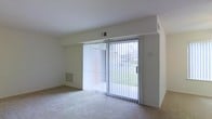 This single-floor residence offers a clean and functional layout designed for comfortable everyday living. The main living area is finished with wall-to-wall carpeting that adds warmth underfoot, while smooth white walls and a flat ceiling create a bright, neutral backdrop throughout the space. A standout feature of the living area is the full-width sliding glass door, dressed with vertical blinds, that floods the room with natural light and provides direct views of a lush green lawn outside. The door also offers convenient access to the outdoor area, seamlessly connecting interior living with the exterior environment. A floor-level baseboard vent is integrated into the wall beneath the sliding door, contributing to the home's climate control system. Overhead, a ceiling vent ensures consistent air circulation throughout the room. The overall ambiance is light, airy, and move-in ready, with a simple and uncluttered design that provides a versatile canvas for a variety of interior styles. The single-story layout ensures easy accessibility, making it a practical and welcoming living environment.