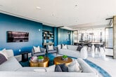 This contemporary single-floor space showcases a thoughtfully designed open-plan layout that seamlessly connects the kitchen, dining, and living areas. Floor-to-ceiling glass walls and sliding doors flood the interior with abundant natural light while offering sweeping outdoor views of a spacious terrace beyond.

The kitchen is anchored by a substantial dark stone island with integrated seating, complemented by sleek, full-height cabinetry in warm neutral tones and light hardwood flooring that extends throughout. The kitchen flows directly into the dining area, where a large rectangular black table is surrounded by matching chairs beneath a striking sculptural brass pendant light fixture — a refined focal point that draws the eye upward.

The living area makes a bold design statement with rich teal feature walls, lush teal-toned area rugs, and a curated arrangement of light grey sofas and accent chairs. Brushed gold cylindrical coffee tables add warmth and contrast, while an open shelving unit and wall-mounted display serve both functional and decorative purposes. Exposed concrete structural columns provide an industrial counterpoint to the polished interior finishes.

Throughout, the design strikes a balance between modern minimalism and vibrant character, creating a space that is equally suited to entertaining and everyday use. The seamless indoor-outdoor connection further enhances the expansive, airy ambiance of this well-appointed space.