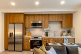 Located at 1350 S Bascom Ave in San Jose, California, this thoughtfully designed studio offers a modern open-concept layout that efficiently combines living, sleeping, and dining areas into one cohesive space. Light wood-tone flooring flows throughout, complemented by recessed lighting and a clean, contemporary color palette that gives the interior a bright, airy feel. Large windows invite natural light and provide views of the building's attractively landscaped courtyard below.

The kitchen is a standout feature, outfitted with warm wood-grain cabinetry extending to the ceiling, stainless steel appliances including a full-size refrigerator, electric range, microwave, and dishwasher, along with light quartz countertops and a tiled backsplash. The well-appointed bathroom includes a full soaking tub and shower combination, wood-front vanity with a white countertop, and neutral tile flooring, creating a calm and spa-like atmosphere. A dedicated closet and hallway provide practical storage and circulation.

The building exterior showcases a contemporary multi-story design with a mix of white paneling and warm wood cladding. Outdoor communal areas feature manicured planters, seating, and lush greenery — offering a welcoming urban retreat in the heart of San Jose.