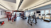 This expansive fitness center offers a thoroughly equipped, single-level layout designed to accommodate a wide variety of training disciplines. The space is organized into distinct functional zones, each tailored to a specific workout focus. The strength training area is anchored by prominent red Rogue power racks and multi-functional cable systems, accompanied by a full set of barbells, weight plates, and tiered dumbbell racks ranging across multiple weight increments. Adjustable benches and a heavy-duty punching bag round out the free weights section.

A dedicated cardio zone features treadmills, stair climbers, stationary bikes, and elliptical trainers, arranged along windowed walls to maximize natural light and provide views of the surrounding exterior. A bold navy accent wall adds a dynamic visual contrast to the otherwise clean, white interior palette.

Tray ceilings with recessed lighting contribute to a polished, modern aesthetic throughout, while durable rubber and carpet flooring provide appropriate surface coverage for varied training activities. Large windows along multiple walls flood the interior with daylight. Additional amenities include a water fountain, wall-mounted fans, a digital clock display, and storage shelving, ensuring both functionality and comfort throughout the facility.