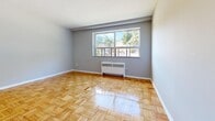 Located at 1712 Main St W in Hamilton's vibrant west end, this one-bedroom, one-bathroom unit offers a practical and well-organized layout across a single floor. The space is defined by warm parquet hardwood flooring throughout the living, bedroom, and hallway areas, lending a classic character to the interior.

The open kitchen and living area flows seamlessly from the entry. The kitchen features white cabinetry, a white range, and light-toned tile flooring, providing a clean and functional cooking space. The adjacent living area benefits from a large window that invites generous natural light and frames a pleasant view of the tree-lined surroundings. A radiator unit ensures comfortable heating.

The bedroom is a well-proportioned, quiet retreat featuring the same warm parquet flooring, a built-in closet with bifold doors, and a window overlooking the greenery outside. Its proximity to the bathroom adds everyday convenience.

The bathroom is neatly finished with floor-to-ceiling white subway-style tiles, a full bathtub with a shower, and a clean white vanity, creating a crisp, functional space.

Throughout the unit, neutral grey walls and white trim contribute to a bright, cohesive aesthetic. This unit is well-suited for those seeking a compact, efficiently designed space in a convenient Hamilton location.