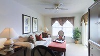 This single-floor space showcases a comfortable and thoughtfully arranged living area that blends classic design with warm, inviting elements. The living room is anchored by a plush white sofa adorned with decorative pillows in rich earth tones, complemented by a deep red upholstered ottoman that doubles as a coffee table. A ceiling fan with a brass finish is centrally mounted on the textured ceiling, promoting airflow and adding a traditional touch to the room.

Natural light filters in through a pair of windows dressed with elegant dark brown valances and sheer white panels, creating a soft and welcoming ambiance. The neutral-toned carpeted flooring enhances the warmth of the space, while framed artwork in dark borders lines the walls, contributing a curated, homey feel. Lush potted plants placed near the window introduce a refreshing natural element. A classic table lamp provides supplemental lighting, rounding out the functional yet stylish arrangement. The overall atmosphere is calm and lived-in, suited for relaxed everyday living.