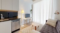 Located at 2061 Rue Stanley in the heart of downtown Montréal, this 2-bedroom, 1-bathroom unit offers a clean, minimalist aesthetic defined by a crisp white palette and warm light wood flooring throughout. The open-plan living and kitchen area forms the heart of the space, where floor-to-ceiling sheer curtains filter abundant natural light, creating an airy, welcoming ambiance. The kitchen is equipped with dark countertops and stainless steel appliances, including a dishwasher, offering a sleek contrast to the bright surroundings. The living area is compact yet functional, with room for a seating arrangement and a small workspace. The two bedrooms maintain the unit's consistent minimalist character, featuring light wood floors, white walls, and large windows that invite natural light and urban views of the surrounding Montréal cityscape. The primary bedroom offers a comfortable footprint, while the second bedroom provides an equally versatile space. The bathroom is finished with large-format light grey tiles on both the floors and walls, a glass-enclosed shower, and modern fixtures, lending a clean and contemporary feel. A hallway connects the rooms efficiently, completing this well-organized and thoughtfully designed urban residence.