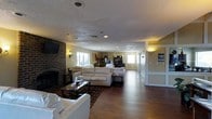 This single-floor space presents an open-concept layout that seamlessly connects the living, dining, and kitchen areas into one expansive, cohesive environment. The living area is anchored by a prominent floor-to-ceiling brick fireplace on the left wall, which serves as a strong architectural focal point. A wall-mounted flat-screen television is positioned above the fireplace, complemented by decorative sconce lighting that adds warmth to the surrounding yellow-toned walls.

The seating area features multiple cream-colored sofas arranged around a dark wood and glass coffee table, set atop a patterned area rug over rich hardwood flooring that extends throughout the space. Recessed lighting in the textured ceiling provides consistent illumination across the open floor plan.

Toward the rear, the space transitions into a dining and kitchen zone, where pendant lighting hangs above what appears to be a counter or island, and dark cabinetry lines the back wall. Windows along the perimeter allow natural light to filter in. A hallway visible to the right suggests additional rooms or access points, contributing to the overall functional and well-connected layout of this inviting space.