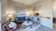 Located at 4816 McMahon Blvd NW in Albuquerque, New Mexico, this one-bedroom, one-bathroom unit offers a smartly designed single-floor layout that maximizes every square foot. The open-concept living area features light wood-look flooring, neutral warm-toned walls, and a seamless flow between the living room and kitchen. The kitchen includes a bar-height counter with seating for two, a stainless steel sink, and a mirrored backsplash that adds depth to the space. A large sliding glass door in the living area opens to a private balcony overlooking lush tree canopies, filling the interior with natural light and outdoor views. The bedroom is generously proportioned, accommodating a queen-sized bed with ease, and includes a mirrored sliding-door closet that enhances the sense of space. A round wall mirror and natural light from a window further brighten the room. The bathroom features a full tub-shower combination with clean white tile, wood-look flooring, and crisp, bright finishes throughout. The unit's cohesive neutral palette, modern fixtures, and thoughtful layout make it a well-appointed and functional living space.