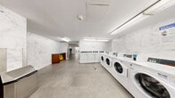 Located at 1959-1999 Marine Drive in North Vancouver, BC, this well-appointed shared laundry facility offers a generous, single-floor layout designed to accommodate a high volume of residents with ease. The space is cleanly finished throughout, with marble-effect wall tiling lining the laundry areas, large-format grey floor tiles, and bright fluorescent lighting that ensures excellent visibility across the entire facility.

The main laundry area is expansive, housing an extensive row of front-loading washing machines and top-loading dryers along one wall, providing ample capacity for concurrent use. A utility sink with cabinetry offers additional convenience for hand-washing or pre-treatment needs. The marble-patterned tile walls lend the laundry area a polished, upscale appearance that elevates the overall aesthetic of the space.

Connecting to the laundry room is a comfortable waiting lounge area, furnished with plush brown leather armchairs arranged to encourage relaxed waiting. An informational kiosk and bulletin board are conveniently positioned near the entrance, keeping residents informed of relevant notices. Vending machines are also available, adding a practical amenity for visitors making use of the facility.

The overall ambiance is clean, functional, and well-maintained, reflecting a commitment to resident comfort and convenience throughout this shared community space.