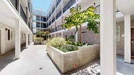 Located at 742 S Westlake Ave in the Westlake neighborhood of Los Angeles, this open-air courtyard serves as a central communal space within a modern multi-story residential building. The courtyard is designed around a long, spacious walkway that runs through the heart of the property, flanked by large concrete raised planters filled with ornamental grasses, succulents, and mature trees that provide natural shade and greenery.

The surrounding building features a clean contemporary aesthetic, with stucco exteriors in neutral tones, white structural columns, and dark metal railings lining the upper-floor balconies. Multiple levels of corridor-access units overlook the courtyard, creating an open, airy atmosphere enhanced by abundant natural sunlight streaming in from above.

At ground level, the courtyard offers casual seating areas furnished with colorful tables and chairs in vibrant shades of green, yellow, and purple, contributing a lively and welcoming character to the space. The wide, smooth concrete walkway provides ample room for movement and social interaction throughout the corridor.

The open-sky design allows natural light to flood the space throughout the day, while the layered landscaping softens the architectural hardscape and adds a sense of vitality. The overall ambiance is bright, modern, and community-oriented, offering residents an inviting outdoor environment at the center of the building.