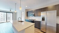 This contemporary single-floor residence showcases a thoughtfully designed open-concept layout with modern finishes throughout. The kitchen serves as the centerpiece, featuring a large island with a quartz countertop, an undermount sink, and stylish globe pendant lighting overhead. Dark wood-grain cabinetry reaches to the ceiling, complemented by a classic white subway tile backsplash and a full suite of stainless steel appliances, including a side-by-side refrigerator with ice and water dispenser, an electric range, and a built-in microwave. Light wood-tone flooring flows throughout the main living areas, adding warmth to the neutral color palette. The bedroom offers plush carpeting, a ceiling fan, and expansive floor-to-ceiling sliding glass doors that flood the space with natural light and offer views of the surrounding urban environment. An additional room with matching large windows and hardwood-style flooring provides flexible living options. The bathroom features light wood cabinetry, a white quartz vanity, and elegant marble-look tile flooring. A dedicated laundry room includes a full-size washer and dryer alongside ample upper cabinetry for storage. The overall ambiance is clean, bright, and move-in ready, reflecting a polished modern aesthetic.