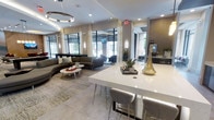 This elegantly designed single-floor space exudes a contemporary and upscale ambiance, blending refined aesthetics with functional comfort. The open-plan layout is anchored by a sleek white quartz countertop bar area, complemented by modern gray upholstered barstools and accented with decorative gold and green sculptural pieces that add an artistic touch.

The lounge area features a generous curved sectional sofa in a rich charcoal tone, paired with a round marble-top coffee table and layered area rugs that define the seating zone. Soft, neutral tones throughout — from the large-format porcelain tile flooring to the cream-colored drapery — create a calm and cohesive aesthetic.

Floor-to-ceiling windows with black steel framing line the perimeter, flooding the interior with abundant natural light and offering views of the surrounding exterior landscape. The ceiling draws the eye upward, adorned with a striking circular pendant light fixture and recessed lighting that adds warmth and depth to the space.

Textural contrast is introduced through exposed brick accent walls and warm wood-toned elements, balancing the otherwise cool, modern palette. Wall-mounted sconces provide soft ambient lighting, further enhancing the polished yet welcoming atmosphere of this thoughtfully curated communal space.